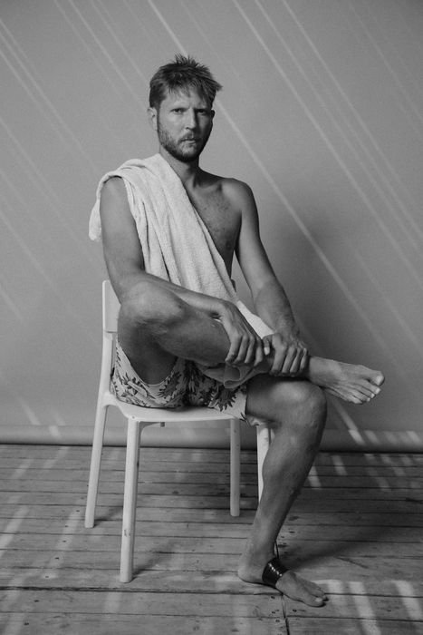 Black and white portrait of a man sitting on a plastic chair against a bare wall, a towel draped over one shoulder, swim shorts, bare feet, one ankle taped.