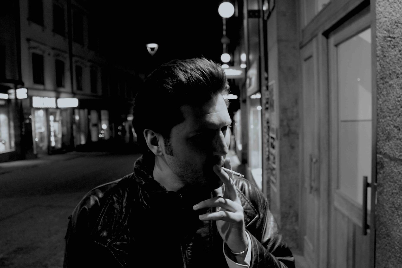 Black and white photograph of a man in a leather jacket smoking a cigarette on a city street at night, shopfronts lit behind him.